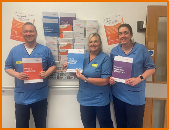 Three members of hospital staff stand infront of a wall-mount patient information dispenser which is full of Myeloma UK patient information. The staff are smiling and holding examples of the patient information.